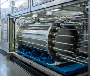 A large industrial machine features a cylindrical metal tank with numerous rods extending from its front, mounted on a blue base. Above it, a clear enclosure reveals complex piping and machinery, suggesting a sophisticated manufacturing or processing system.