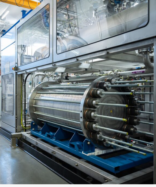 A large industrial machine features a cylindrical metal tank with numerous rods extending from its front, mounted on a blue base. Above it, a clear enclosure reveals complex piping and machinery, suggesting a sophisticated manufacturing or processing system.