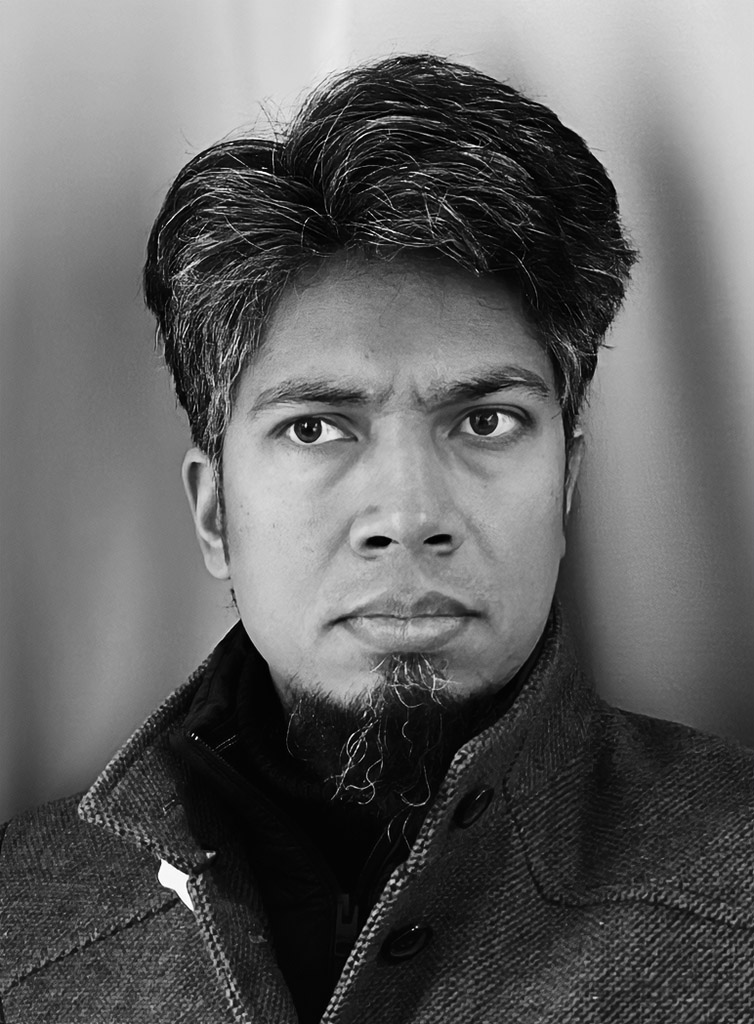 A black and white headshot shows a man with dark, slightly disheveled hair and a small goatee. He is wearing a collared jacket and a dark turtleneck, looking directly at the camera with a neutral expression.