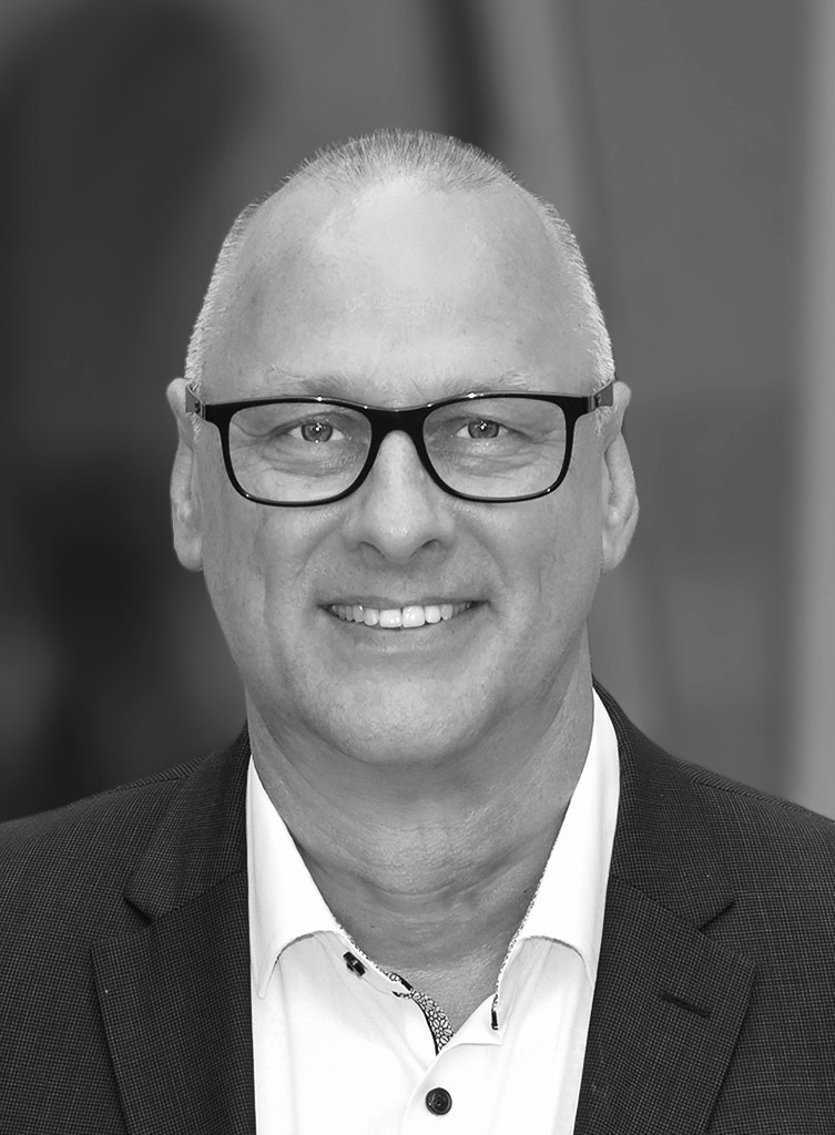 A black and white headshot shows a smiling man with a shaved head and glasses. He is wearing a suit jacket and a white collared shirt with the top button undone.