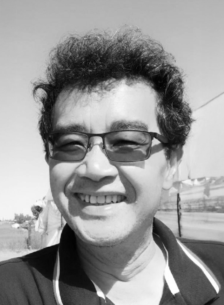 A black and white close-up shows a man with messy dark hair and glasses, smiling broadly at the camera. He is wearing a collared shirt with stripes and appears to be outdoors under a bright sky.