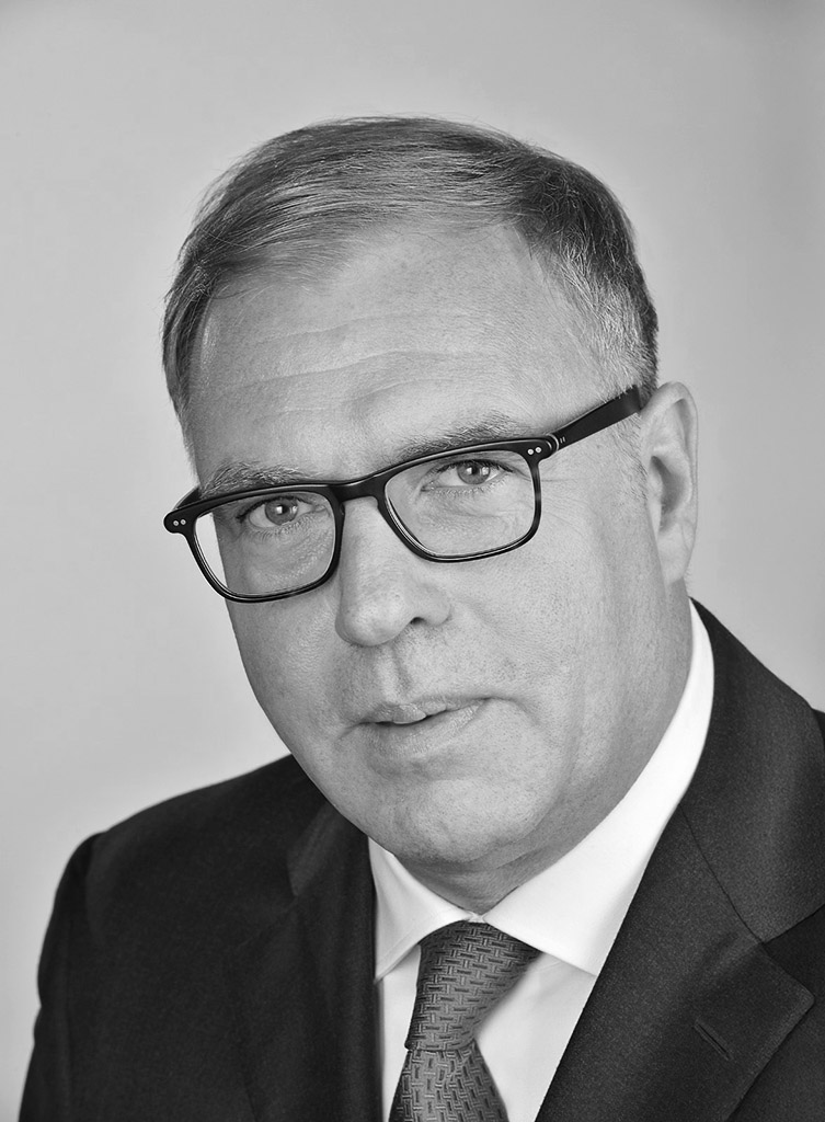 A black and white headshot shows a man wearing glasses and a suit with a patterned tie. He has short, neatly combed hair and is looking directly at the camera with a serious expression.